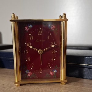 Puede incluir: Un reloj de viaje rectangular dorado con esfera burdeos. La esfera presenta detalles florales y la marca "Bucherer". El reloj tiene una cubierta de vidrio y un marco dorado. El reloj está sobre una superficie de madera.