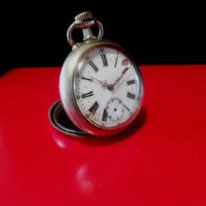 May include: A silver pocket watch with Roman numeral markings on the face. The watch has a white face with black hands and a small second hand dial at the bottom.
