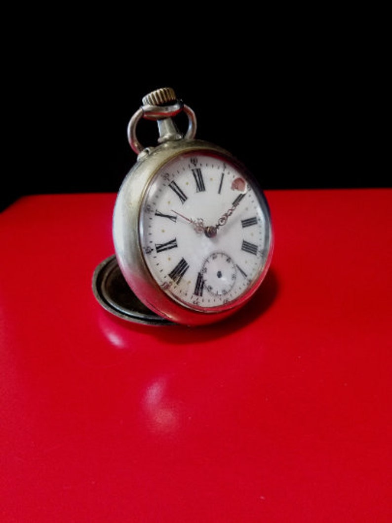 May include: A silver pocket watch with Roman numeral markings on the face. The watch has a white face with black hands and a small second hand dial at the bottom.