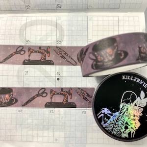 May include: Purple washi tape featuring sewing-themed illustrations, including scissors and sewing machines. A black round sticker with the text "KILLERVILLE" and a holographic design is also visible. The tape is on a calendar.
