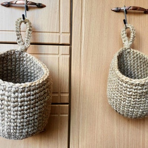 Puede incluir: Dos cestas colgantes de crochet marrón con asas de cuerda, colgadas de ganchos en una pared de madera.