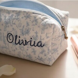 May include: A white cosmetic bag with a blue floral pattern and the name "Olivia" embroidered in navy blue. The bag has a light blue gingham lining and a gold zipper.
