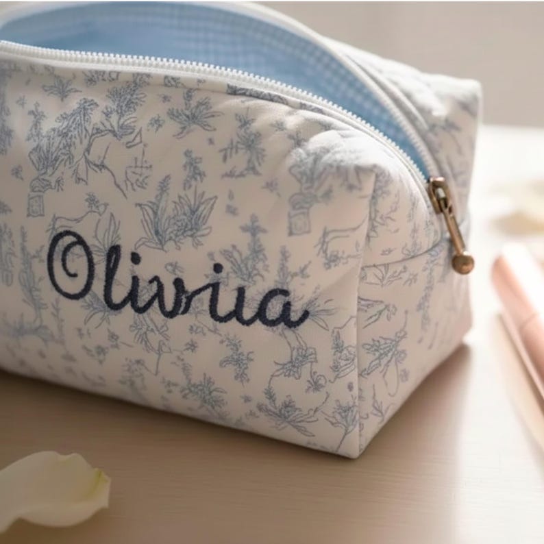 May include: A white cosmetic bag with a blue floral pattern and the name "Olivia" embroidered in navy blue. The bag has a light blue gingham lining and a gold zipper.