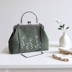 May include: A sage green handbag with a silver clasp and handle. The bag has a white floral embroidery design. A silver chain strap hangs from the bag. The bag is on a white surface.