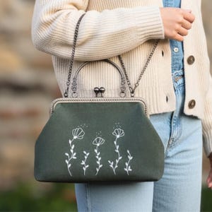 May include: A dark green handbag with a silver clasp and chain strap. The bag features white floral embroidery. The bag is held by a person wearing a cream cardigan and blue jeans.