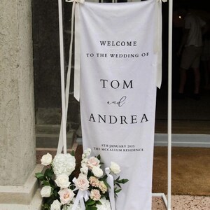 Op de afbeelding: Witte bruiloft welkom bord met zwarte tekst. Het bord staat "Welkom op de bruiloft van Tom en Andrea, 8 januari 2025, The McCallum Residence".