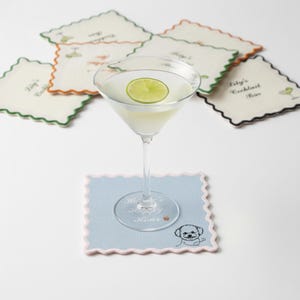 May include: A martini glass with a lime slice sits on a light blue coaster with a scalloped edge and a dog illustration. Several coasters with scalloped edges and text are in the background. The coasters have different colored edges.