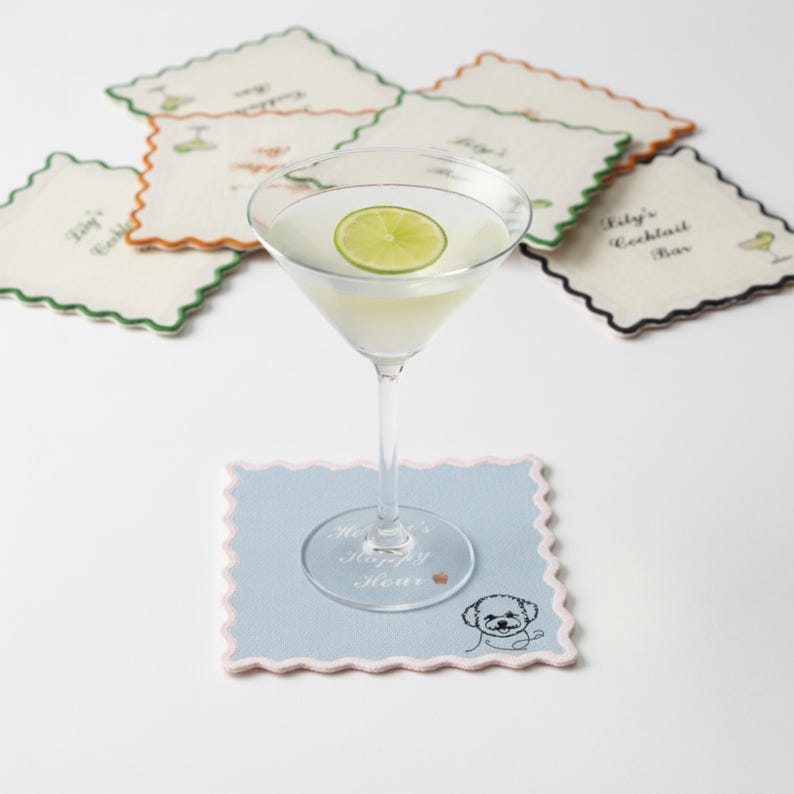 May include: A martini glass with a lime slice sits on a light blue coaster with a scalloped edge and a dog illustration. Several coasters with scalloped edges and text are in the background. The coasters have different colored edges.