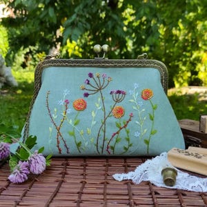 Pu&ograve; includere: Una pochette azzurra con chiusura e cornice in metallo. La pochette &egrave; ricamata con fiori e steli colorati. Lo sfondo &egrave; un verde sfocato, che suggerisce un ambiente esterno. La pochette &egrave; appoggiata su una superficie di legno.
