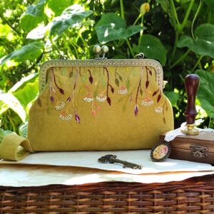 Pu&ograve; includere: Una pochette color senape con chiusura in metallo decorativa. La pochette &egrave; ricamata con un motivo floreale nei toni del verde, viola e bianco. Una chiave e un piccolo ciondolo sono accanto alla pochette.
