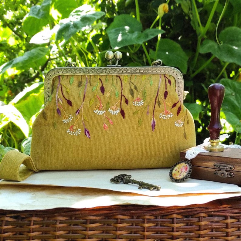 Pu&ograve; includere: Una pochette color senape con chiusura in metallo decorativa. La pochette &egrave; ricamata con un motivo floreale nei toni del verde, viola e bianco. Una chiave e un piccolo ciondolo sono accanto alla pochette.