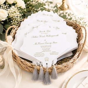 May include: A stack of white scalloped-edge cardstock fans in a woven basket, featuring gold foil text that reads Save the Date for the wedding of Arthur Woods Hollingsworth & McKenzie Truong Starr, with grey tassels attached to the bottom of each fan.