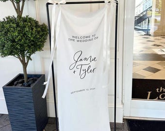 May include: White wedding welcome sign with black text. The sign reads "Welcome to the wedding of Jamie + Tyler, September 13, 2024".