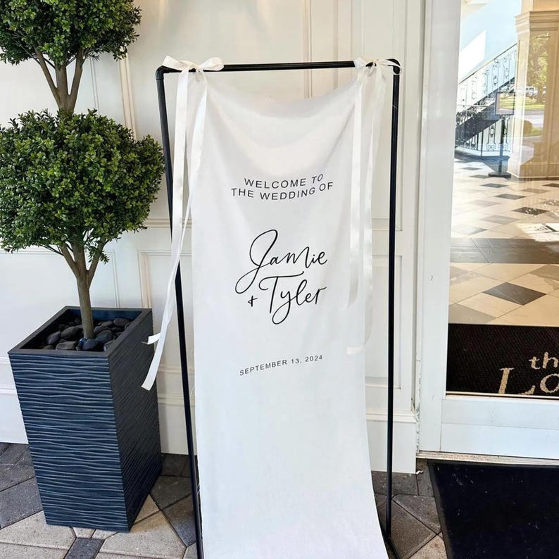 Op de afbeelding: Witte bruiloft welkomstbord met zwarte tekst. Het bord staat "Welkom op de bruiloft van Jamie + Tyler, 13 september 2024".
