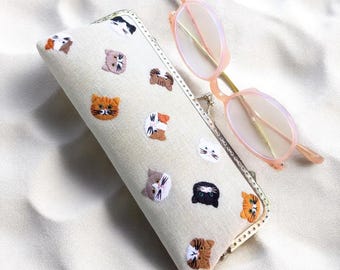 Op de afbeelding: Een beige brillenkoker geborduurd met kattengezichten in oranje, bruin, zwart en wit. Een lichtroze bril met een gouden montuur ligt naast de koker.