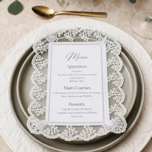 May include: A formal place setting with a menu card. The menu card is white with a decorative lace border and features the word "Menu" at the top. The menu lists appetizers, main courses, and desserts. A gold spoon is in the background.