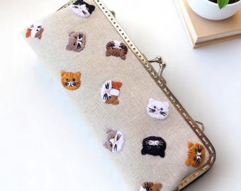 May include: A beige fabric clutch with a metal clasp, adorned with embroidered cat faces in various colors. The cats have different expressions and patterns, creating a whimsical design. The clutch is rectangular and appears to be handmade.