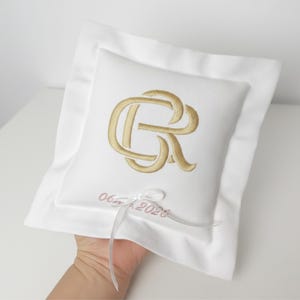 May include: A white ring pillow with a gold embroidered monogram and the date "06.06.2023" in pink. The pillow has a square shape with a ruffled border and a white ribbon.