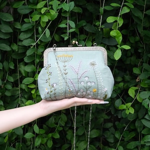May include: A light green clutch with a metal clasp and chain strap. The clutch is embroidered with a floral design in white, yellow, and purple thread. The bag is held in front of a green leafy background.