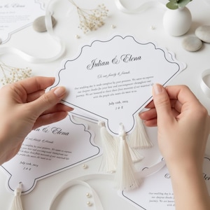 Puede incluir: Invitaciones de boda blancas con borde festoneado y detalles de borlas. Las invitaciones presentan los nombres "Julian & Elena" y los detalles del evento. El diseño incluye una escritura elegante y un borde decorativo.