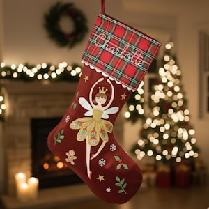 Pode incluir: Meia de Natal bordô com punho xadrez bordado com o nome "Charlotte". A meia apresenta uma fada bailarina bordada com tutu dourado, asas e coroa. Detalhes bordados adicionais incluem estrelas, flocos de neve e galhos.
