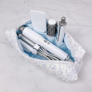 May include: A toiletry bag with a blue and white floral pattern, containing various white and silver hair styling tools. The bag has a blue gingham lining and a zipper closure. Items include a hair dryer, curling iron, and brush.