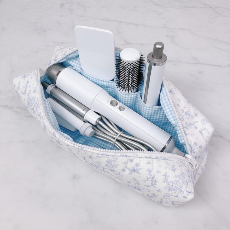 May include: A toiletry bag with a blue and white floral pattern, containing various white and silver hair styling tools. The bag has a blue gingham lining and a zipper closure. Items include a hair dryer, curling iron, and brush.