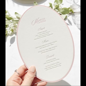 May include: An oval-shaped menu card with a soft pink border and elegant script. The menu lists starters, main courses, and desserts. The card is held by a hand, suggesting a wedding or special event.
