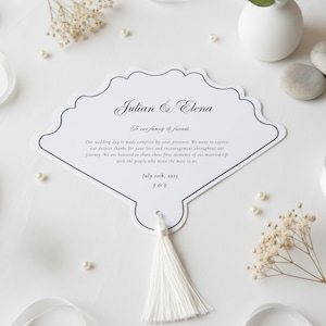May include: A white, fan-shaped wedding invitation with a decorative border and a white tassel. The invitation includes the names "Julian & Elena" and the date "July 12th, 2025". Delicate floral accents and pearls surround the invitation.