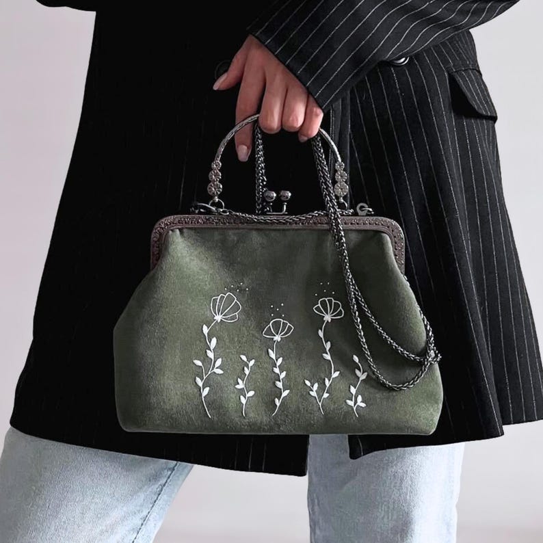May include: A sage green handbag with a floral design. The bag has a metal clasp and handle, with a chain strap. The floral design is white and embroidered on the front of the bag. The bag is held by a person.