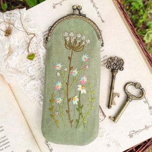 May include: A sage green embroidered glasses case with a floral design. The case has a metal clasp and is decorated with colourful embroidered flowers. Two antique keys are next to the case.