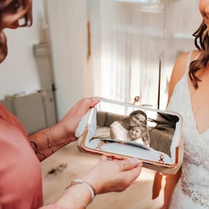 May include: A gold-framed clutch with a clasp reflects a sepia-toned image of a mother and child. The clutch has a light-coloured interior and is held by a person wearing a pink top. The background is a room with a bride in a white dress.