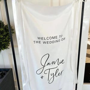 Op de afbeelding: Witte bruiloft welkom bord met zwarte tekst. Het bord zegt "Welkom op de bruiloft van Jamie + Tyler, 13 september 2024".