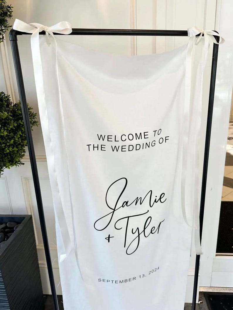 Op de afbeelding: Witte bruiloft welkom bord met zwarte tekst. Het bord zegt "Welkom op de bruiloft van Jamie + Tyler, 13 september 2024".