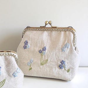May include: Two beige fabric purses with a floral design. The larger purse has a metal clasp and is decorated with embroidered blue flowers and green leaves. The smaller purse is also beige with a metal clasp and embroidered blue flowers.
