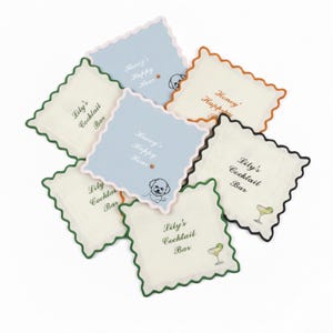 May include: A collection of square coasters with scalloped edges. Some coasters are off-white, others are light blue. Text on the coasters reads "Lily's Cocktail Bar" and "Honey's Happy Hour". A dog illustration is also present.