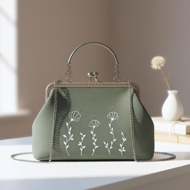 May include: A sage green handbag with a silver metal frame and chain strap. The bag has a white floral design. The handle is a curved silver metal bar. The bag is on a white surface.