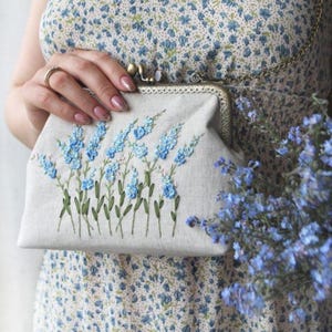 May include: A beige fabric clutch bag featuring an embroidered design of light blue flowers with green stems. The bag has a metallic frame with a clasp closure and a thin gold-toned chain strap. The fabric has a textured, natural appearance.