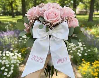 May include: A bouquet of pink roses and other flowers, accented with green leaves and baby's breath. A large white satin bow is tied around the stems, with "2025" and "HALEY" printed in pink.