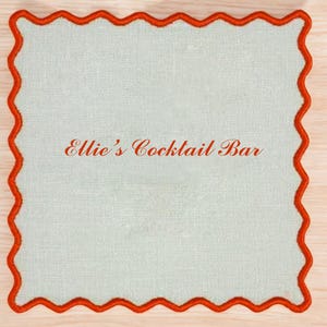 May include: A white square coaster with a red scalloped border. The text "Ellie's Cocktail Bar" is printed in red on the coaster.