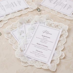 May include: White menu cards with a decorative lace border. The cards display menu items for appetizers, main courses, and desserts. The cards are tied together with a white ribbon. The background is a light beige.