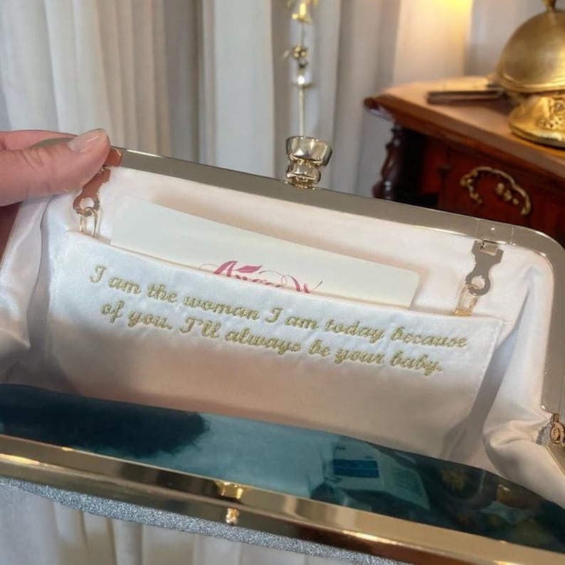 May include: A silver glitter clutch with a gold frame and clasp. The interior features a white satin lining with gold embroidered text that reads, "I am the woman I am today because of you. I'll always be your baby."