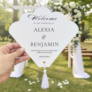 May include: A white fan-shaped wedding programme card with black text that reads Welcome to the wedding of ALEXIA & BENJAMIN, Sont heureux de vous accueillir pour célébrer leur mariage, and the date 20.09.2025, featuring a white tassel at the bottom.