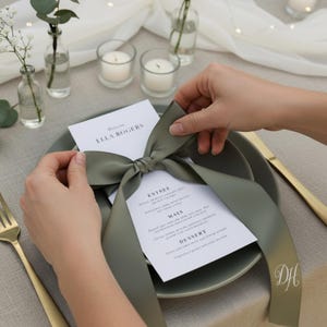 May include: A table setting featuring a sage green plate, menu, and a large satin ribbon tied in a bow. The menu includes the text "Welcome Ella Rogers" and a list of courses. A gold fork and knife are visible.