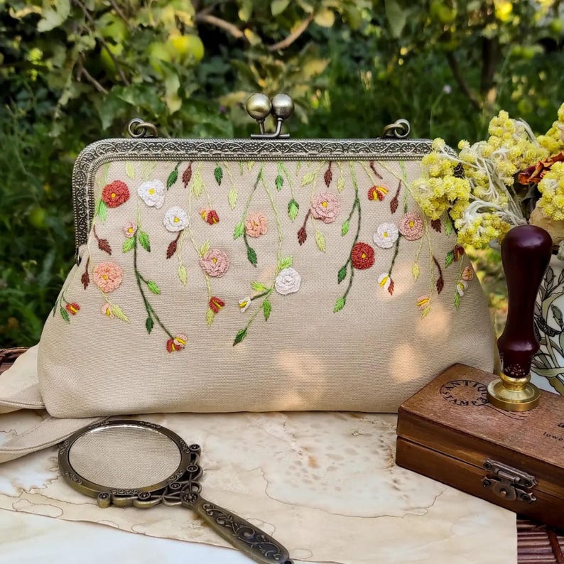 Pu&ograve; includere: Pochette beige con chiusura in metallo e ricami floreali rossi, rosa e bianchi. I fiori ricamati scendono a cascata sulla parte anteriore della borsa. Sono visibili anche uno specchio a mano in stile vintage e una scatola di legno.