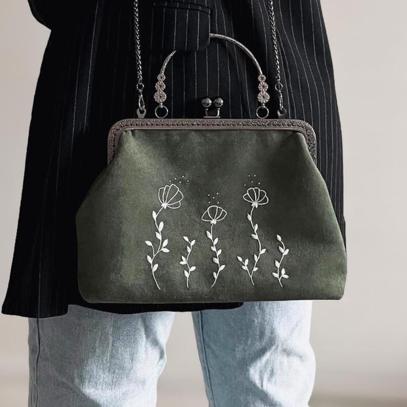 May include: A dark green handbag with a silver clasp and chain strap. The bag features a floral design in white embroidery. The bag is held by a person wearing a black blazer and blue jeans.