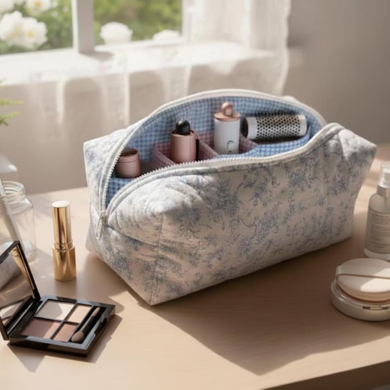 May include: A quilted cosmetic bag with a blue floral pattern and a light blue gingham lining. The bag is open, revealing various cosmetic products. A gold lipstick tube and a black eyeshadow palette are in the foreground.