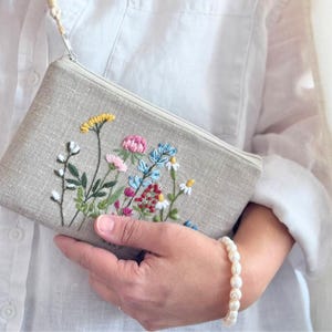 May include: A rectangular, beige linen pouch with a zipper closure and a wrist strap. The pouch is embroidered with a colorful floral design featuring yellow, pink, blue, and white flowers. The pouch is held by a person wearing a white shirt.