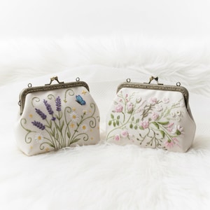Op de afbeelding: Twee crèmekleurige, geborduurde portemonnees met metalen sluitingen. Eén heeft lavendel, madeliefjes en een blauwe vlinder. De andere toont roze rozen en groen. Beide portemonnees liggen op een witte, zachte ondergrond.