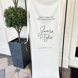 Op de afbeelding: Witte bruiloft welkom bord met zwarte tekst. Het bord staat "Welkom op de bruiloft van Jamie & Tyler, 13 september 2024".